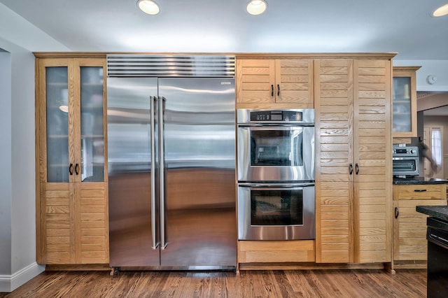 kitchen with appliances with stainless steel finishes, dark stone countertops, light brown cabinetry, and dark hardwood / wood-style flooring