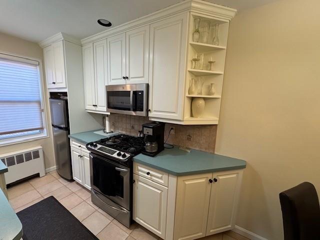 kitchen featuring light tile patterned floors, radiator heating unit, white cabinets, stainless steel appliances, and backsplash