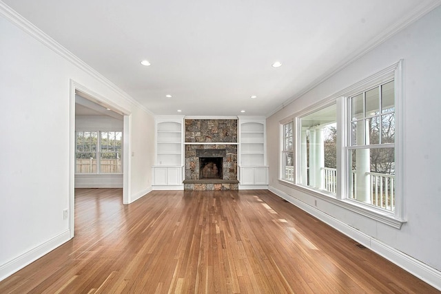 unfurnished living room featuring a stone fireplace, built in shelves, wood finished floors, baseboards, and crown molding