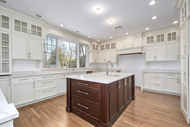 kitchen featuring a center island with sink, visible vents, glass insert cabinets, a sink, and dark brown cabinetry