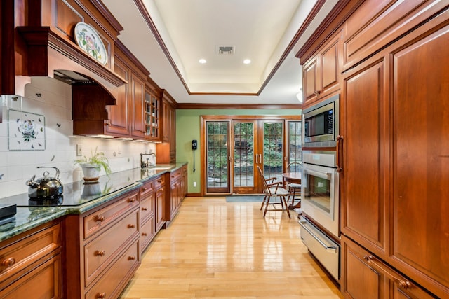 kitchen with light hardwood / wood-style flooring, appliances with stainless steel finishes, backsplash, a raised ceiling, and dark stone counters