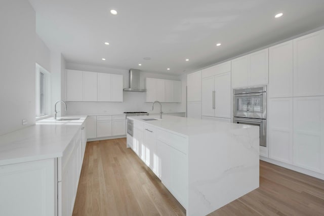 kitchen featuring wall chimney exhaust hood, sink, stainless steel double oven, light hardwood / wood-style floors, and white cabinets