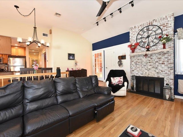 living room featuring vaulted ceiling, a brick fireplace, track lighting, a notable chandelier, and light hardwood / wood-style floors