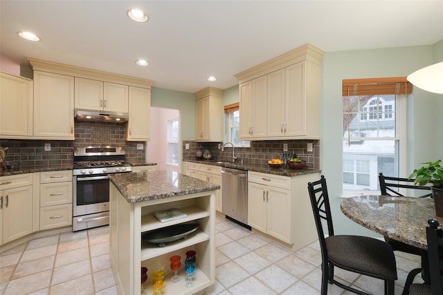 kitchen with appliances with stainless steel finishes, a center island, cream cabinets, under cabinet range hood, and open shelves