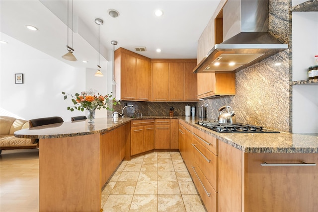 kitchen with stone countertops, hanging light fixtures, kitchen peninsula, gas stovetop, and wall chimney exhaust hood