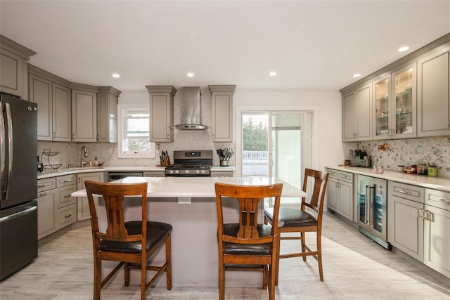 kitchen featuring beverage cooler, gray cabinets, stainless steel appliances, wall chimney range hood, and a kitchen bar