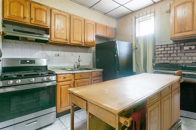 kitchen featuring sink, light tile patterned floors, black refrigerator, stainless steel range with gas stovetop, and decorative backsplash