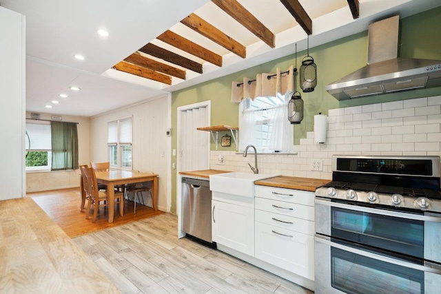 kitchen featuring wall chimney range hood, a sink, wood counters, stainless steel appliances, and tasteful backsplash