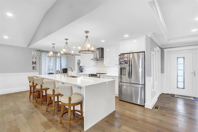 kitchen with white cabinetry, hanging light fixtures, an island with sink, stainless steel appliances, and wall chimney range hood