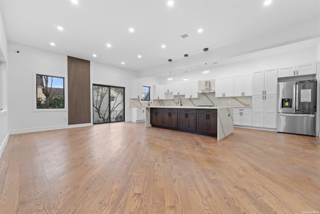 kitchen featuring white cabinets, hanging light fixtures, a center island with sink, stainless steel refrigerator with ice dispenser, and wall chimney range hood