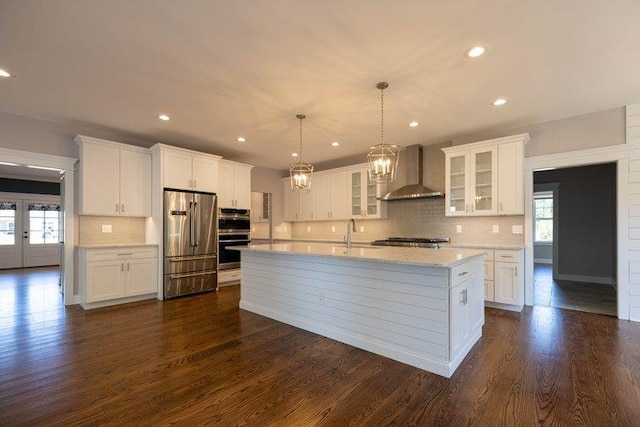 kitchen with appliances with stainless steel finishes, white cabinets, a center island with sink, decorative light fixtures, and wall chimney exhaust hood