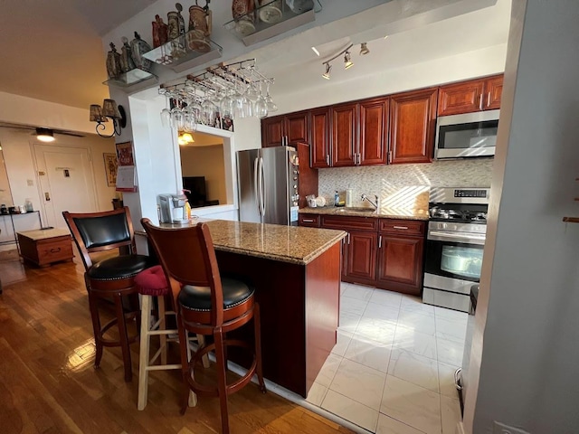 kitchen featuring stainless steel appliances, a sink, backsplash, light stone countertops, and reddish brown cabinets