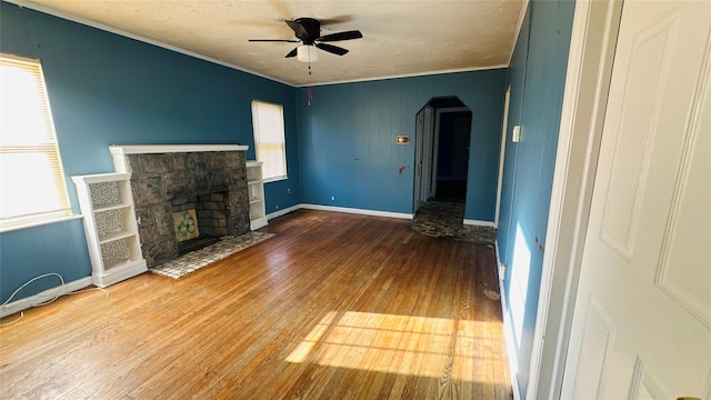 unfurnished living room featuring crown molding, ceiling fan, wood-type flooring, and a stone fireplace