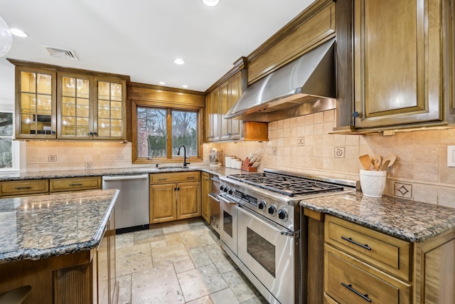 kitchen with stainless steel appliances, stone tile flooring, brown cabinetry, a sink, and wall chimney range hood