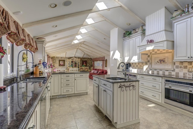 kitchen featuring sink, lofted ceiling with skylight, a center island with sink, dark stone counters, and stainless steel oven