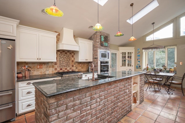 kitchen with hanging light fixtures, dark stone countertops, custom range hood, stainless steel appliances, and a kitchen island with sink