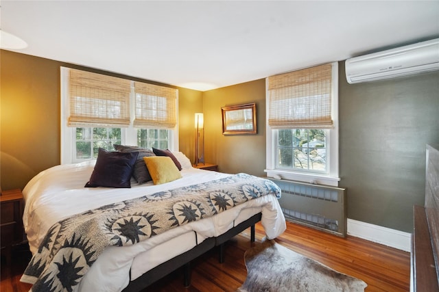 bedroom with a wall unit AC, radiator heating unit, baseboards, and wood finished floors