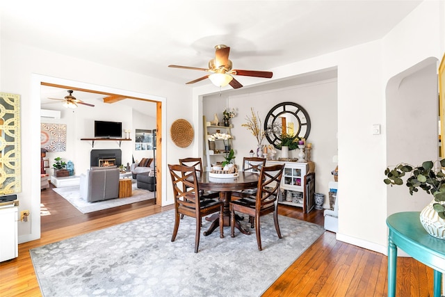 dining area featuring a warm lit fireplace, wood-type flooring, ceiling fan, and an AC wall unit