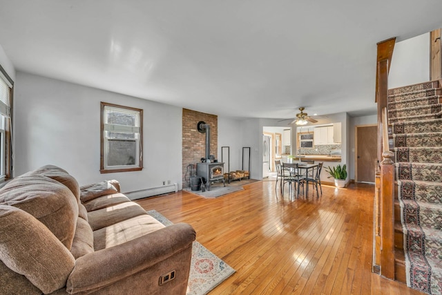 living room with baseboard heating, ceiling fan, a wood stove, and light wood-type flooring