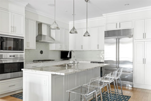 kitchen with wall chimney exhaust hood, built in appliances, light stone countertops, a kitchen island with sink, and white cabinets