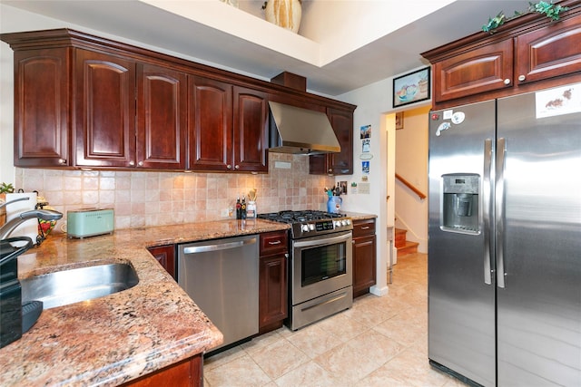 kitchen with light stone counters, stainless steel appliances, a sink, backsplash, and wall chimney exhaust hood