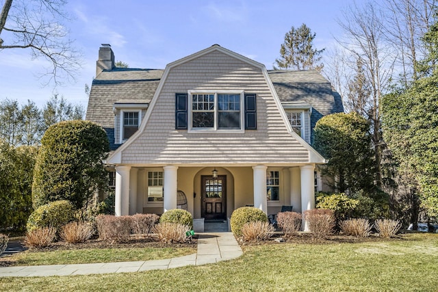 view of front of home featuring a gambrel roof, a chimney, roof with shingles, a front yard, and stucco siding