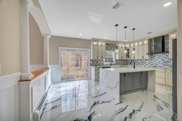 kitchen featuring light stone counters, baseboard heating, an island with sink, and ornate columns