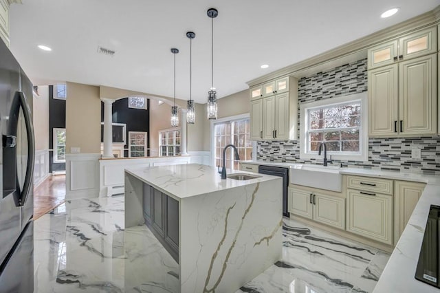 kitchen featuring stainless steel refrigerator with ice dispenser, sink, light stone counters, a kitchen island with sink, and decorative columns