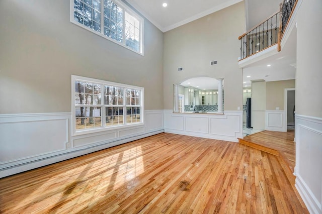 unfurnished living room featuring baseboard heating, crown molding, light hardwood / wood-style floors, and a high ceiling