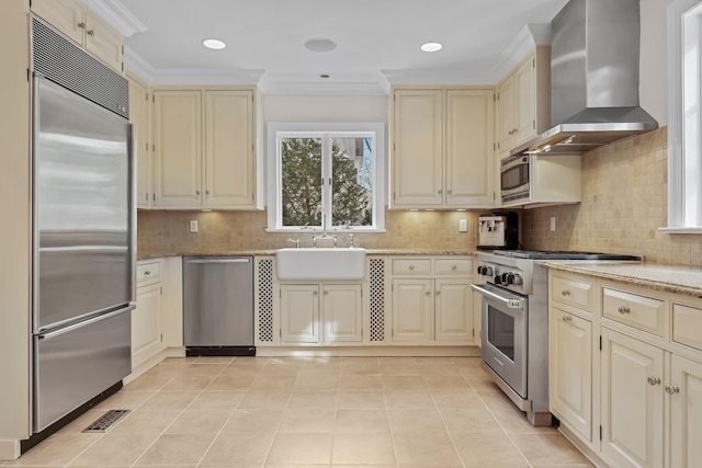 kitchen with wall chimney range hood, built in appliances, sink, and cream cabinets