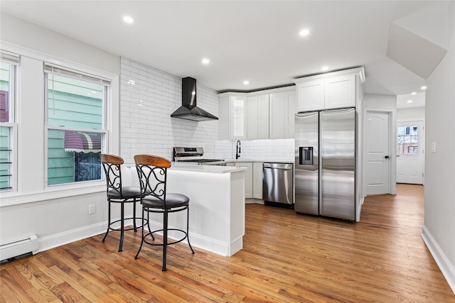 kitchen featuring a baseboard radiator, white cabinets, kitchen peninsula, stainless steel appliances, and wall chimney range hood