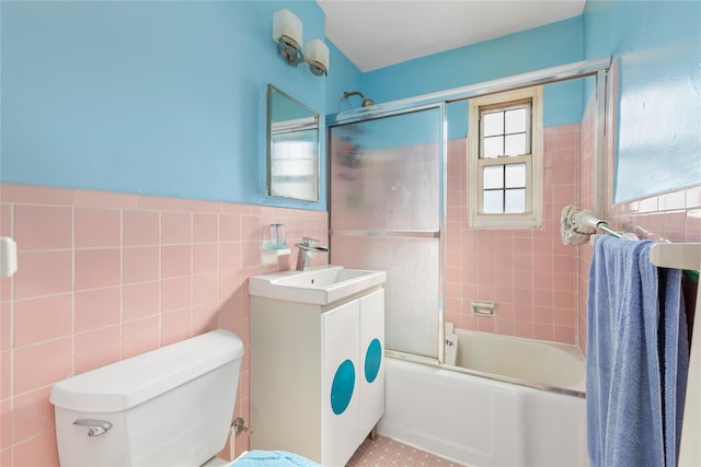 full bathroom featuring toilet, bath / shower combo with glass door, vanity, tile walls, and wainscoting