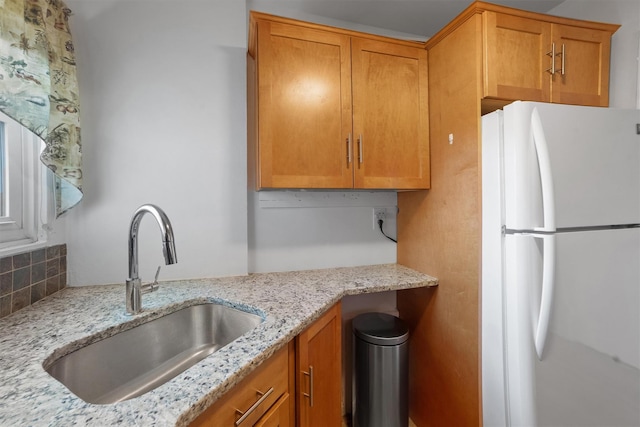 kitchen featuring light stone countertops, a sink, decorative backsplash, freestanding refrigerator, and brown cabinets