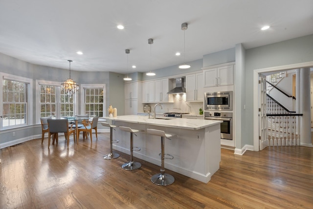 kitchen featuring wall chimney range hood, hanging light fixtures, stainless steel appliances, white cabinets, and a center island with sink