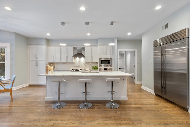 kitchen featuring built in appliances, pendant lighting, wall chimney range hood, a kitchen island with sink, and white cabinets