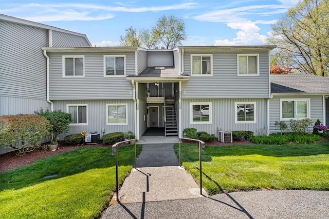 townhome / multi-family property featuring central AC unit, a shingled roof, a front lawn, and stairway