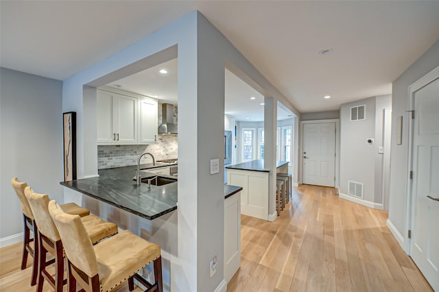 kitchen with dark countertops, visible vents, white cabinetry, a sink, and wall chimney range hood