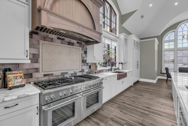 kitchen featuring range with two ovens, light stone counters, custom range hood, white cabinetry, and a sink