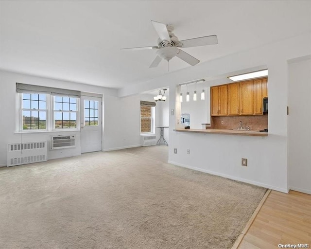 unfurnished living room featuring ceiling fan with notable chandelier, radiator heating unit, sink, light colored carpet, and a wall unit AC