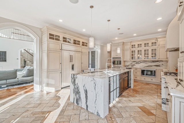 kitchen featuring a center island with sink, glass insert cabinets, built in appliances, hanging light fixtures, and cream cabinets