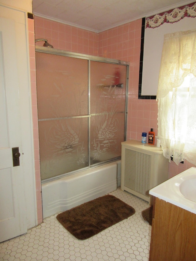 bathroom featuring bath / shower combo with glass door, tile walls, ornamental molding, radiator heating unit, and vanity