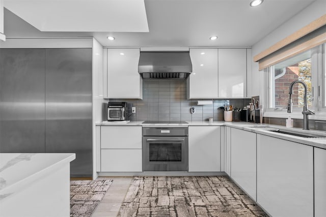 kitchen featuring wall chimney range hood, modern cabinets, a sink, and stainless steel oven