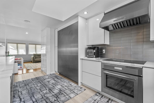 kitchen featuring white cabinets, oven, black electric cooktop, light countertops, and wall chimney range hood