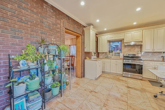 kitchen with brick wall, stainless steel gas range, cream cabinets, under cabinet range hood, and backsplash