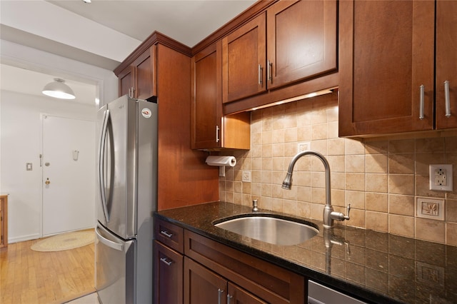 kitchen featuring a sink, freestanding refrigerator, tasteful backsplash, brown cabinetry, and dark stone countertops