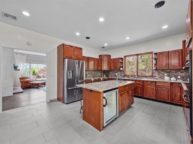kitchen featuring stainless steel fridge, an island with sink, tasteful backsplash, light stone counters, and recessed lighting