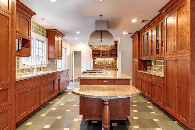 kitchen featuring sink, island range hood, a center island, light stone countertops, and decorative backsplash