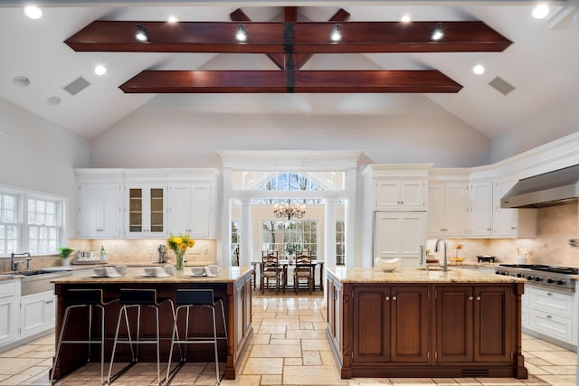 kitchen featuring white cabinetry, stainless steel gas stovetop, a center island with sink, stone tile flooring, and ornate columns