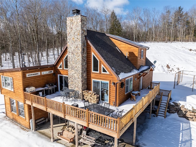 snow covered house featuring a chimney, a deck, and roof with shingles