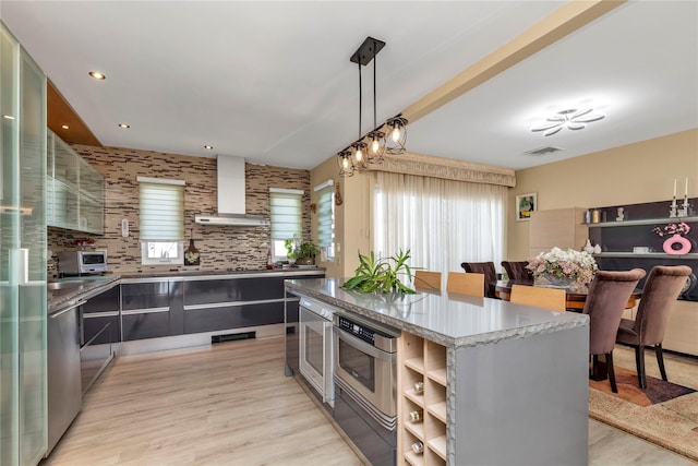 kitchen with visible vents, a kitchen island, glass insert cabinets, light wood-type flooring, and wall chimney range hood